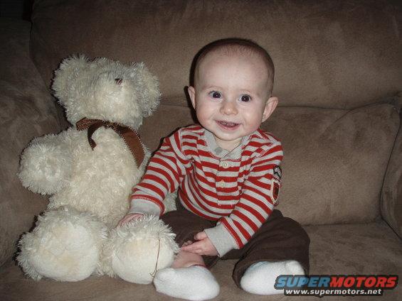 Baby in striped shirt and brown pants sits next to a large white teddy bear on a brown couch