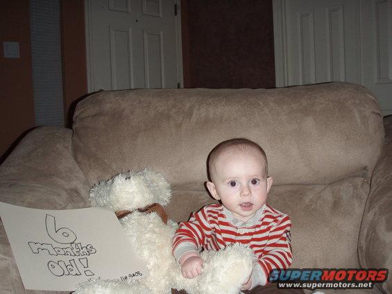 Baby sitting on a couch next to a teddy bear and a sign that says "6 Months Old!"