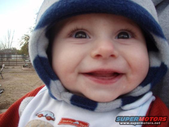 Baby in a blue and gray striped hood, smiling. There's a fence and some trees in the background