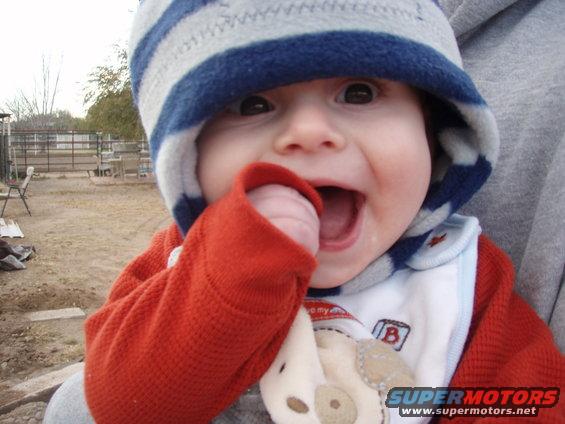 Baby wearing a striped hat and orange sweater, sucking on thumb. Background shows a fence and dry ground