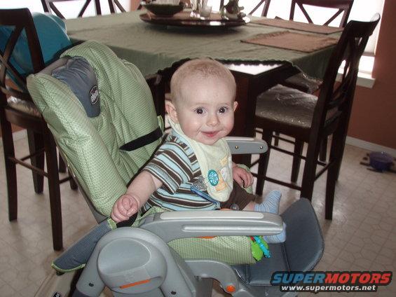 Baby in a green and gray high chair, wearing a striped shirt and bib, with a dining table in the background