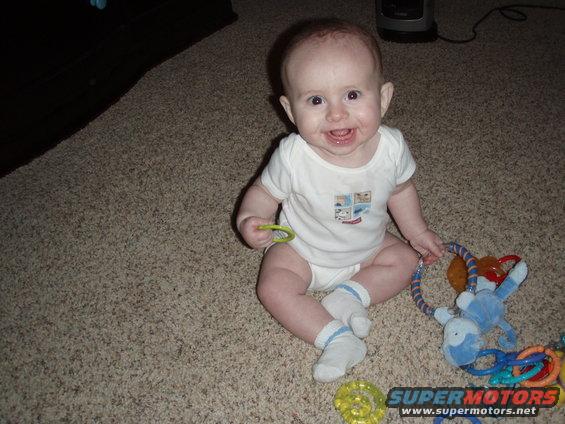 Baby sitting on carpet with toys, Supermotors logo visible