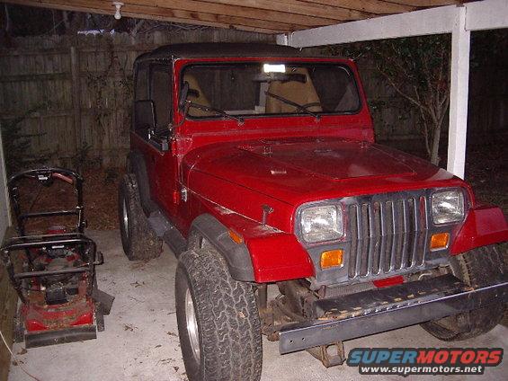 Red Jeep Wrangler parked under a cover next to a lawnmower