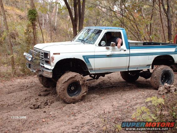 White and blue pickup truck with large tires driving through mud. Chrome bumper and side trim visible