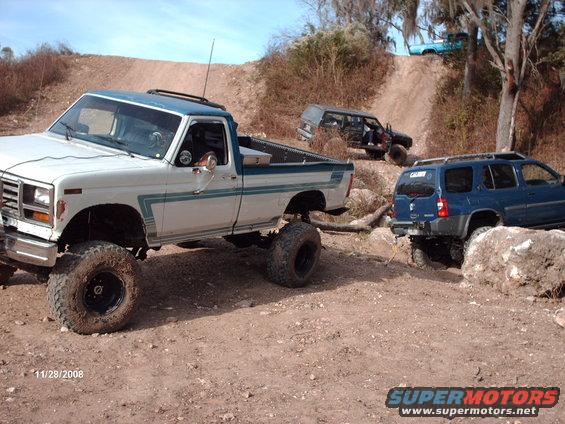 Lifted white and blue pickup truck with off-road tires, blue SUV, and black SUV on a dirt trail