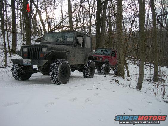 Black Jeep Wrangler with large tires in snow, towing a red Suzuki Samurai