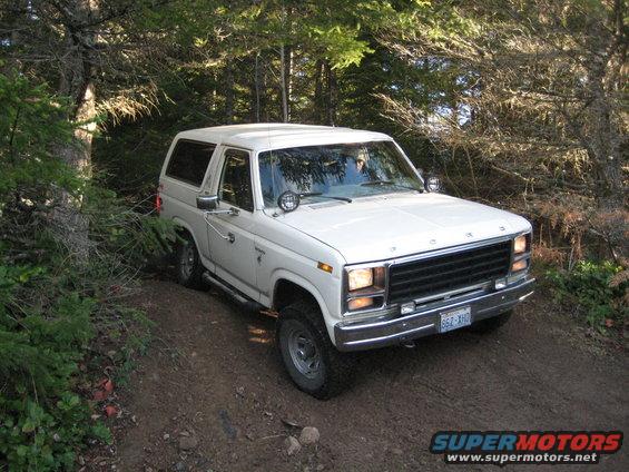 White 1980 Ford Bronco driving on a dirt path in a wooded area