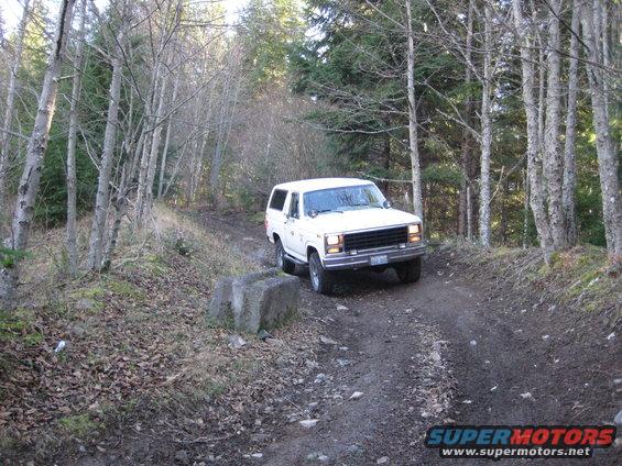 White SUV driving on a dirt road through trees. It looks like a Ford Bronco from the early 80s