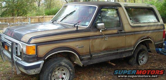 Old Ford Bronco with a brush guard and two-tone paint, sitting in a yard with a flat tire