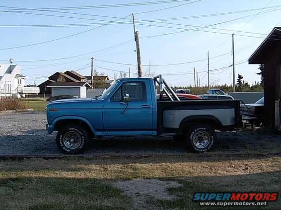 Blue pickup truck with a silver bed, roll bar, and aftermarket wheels. Visible text on tire