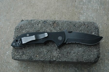 Black folding knife with a partially serrated blade resting on a gray brick
