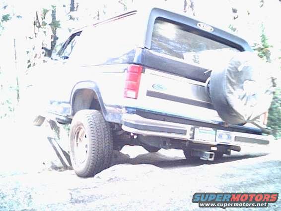 Rear view of a Ford Bronco with a spare tire mounted on the back. It's parked on uneven ground with trees in the background