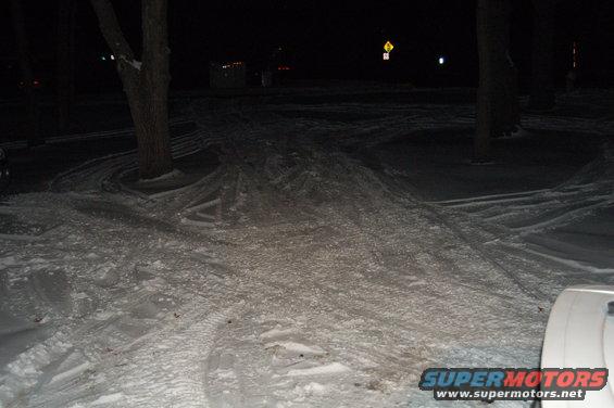 Snowy ground with tire tracks and a partial white vehicle bumper on the right