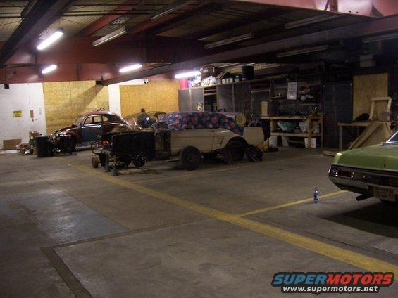 Garage interior with several cars in various states of repair, including a partially covered vehicle and a vintage sedan