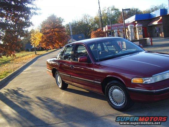 Maroon sedan parked on a road with autumn trees and a gas station in the background