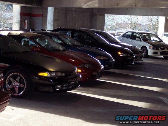 Several Mitsubishi Eclipses in a parking garage, one black, one maroon, one blue, and one dark. A white sedan is parked further back