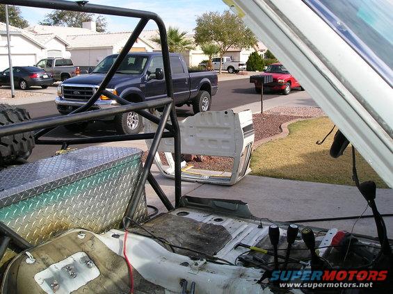Disassembled vehicle interior with roll cage, diamond plate toolbox, and door removed. Pickup truck and red car visible in background