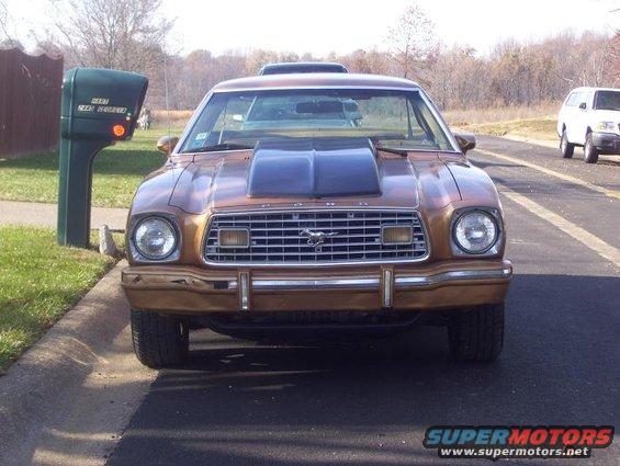 1977 Ford Mustang II with a hood scoop parked curbside, another SUV visible on the road