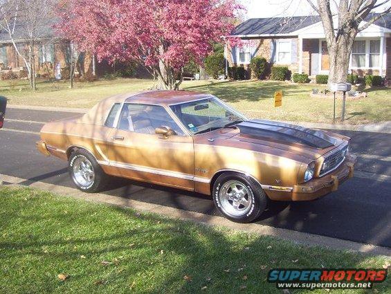 Gold 1977 Ford Mustang II coupe with a black hood scoop parked on a residential street