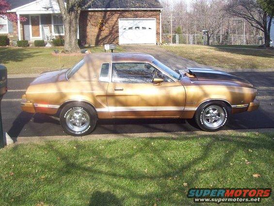 Gold 1977 Ford Mustang II coupe with a brown vinyl top, chrome wheels, and black hood scoop parked on a driveway