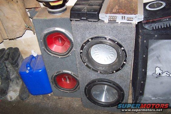Car audio subwoofers in carpeted boxes with amplifiers on top. A blue jerrycan and boots are to the left
