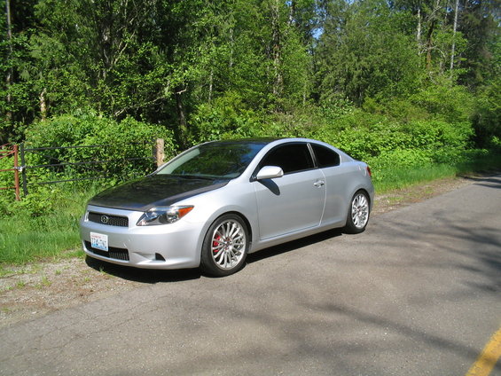 Silver 2005 Scion tC coupe with a black hood and aftermarket wheels parked on a paved road