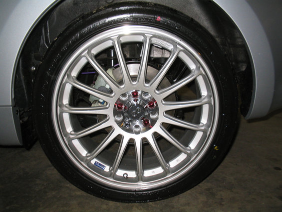 Silver multi-spoke aftermarket wheel with red lug nuts on a wet tire. A blue sticker is visible on the wheel rim