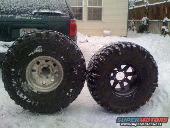 Two large off-road tires, one with a silver rim and one with a black rim, sit in the snow