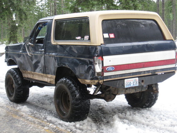 Lifted Ford Bronco with oversized tires, seen from the rear in a snowy wooded area. Has a two-tone paint job and a custom bumper