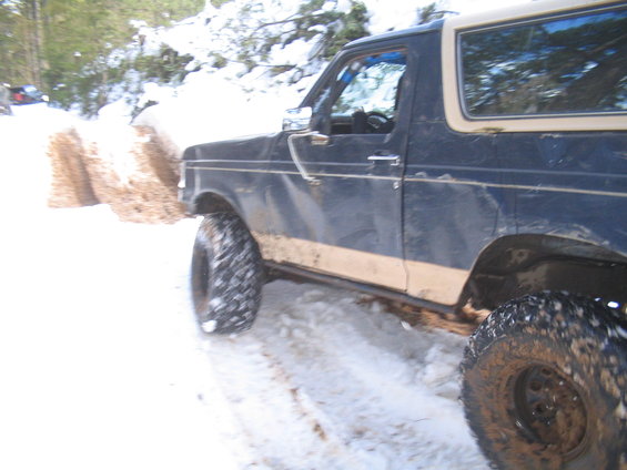 Dark blue Ford Bronco with tan stripe, driving in snow. Lifted suspension and off-road tires visible