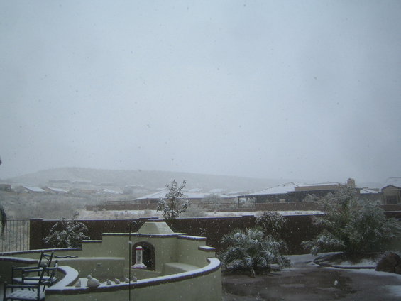 Backyard with snow falling, a patio, chairs, and houses in the background