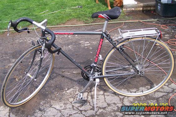 Black Schwinn bicycle with drop handlebars, a rear rack, and tan tires, parked on cracked pavement