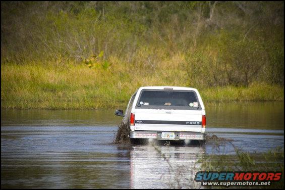 White Ford F-150 driving through shallow water, kicking up mud