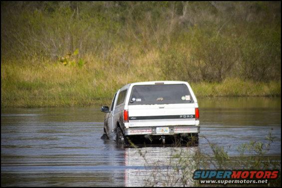 White Ford Bronco driving through water, rear view. Mud splatters on lower body