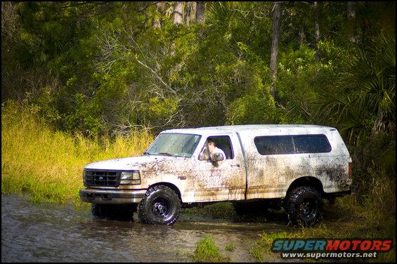 Muddy white pickup truck with a camper shell driving through shallow water near a wooded area