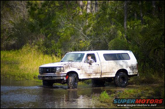 Mud-splattered white Ford F-150 with a camper shell driving through shallow water, front-left tire visible