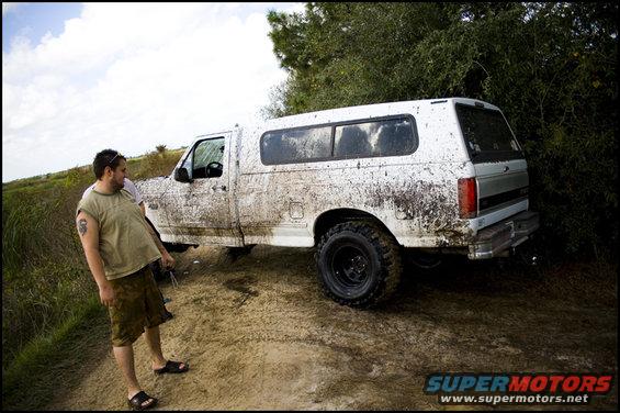 Muddy white pickup truck with a camper shell parked on a dirt road next to some bushes. A man stands to the left of the truck
