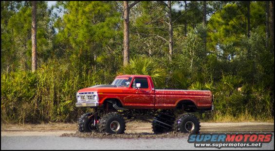 Red lifted pickup truck with large tires driving through mud, side view. Hood scoop visible