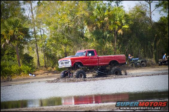 red lifted pickup truck in mud with two ATVs in background