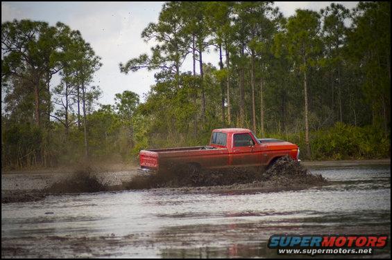Red pickup truck driving through mud, kicking up spray. Trees and water in background