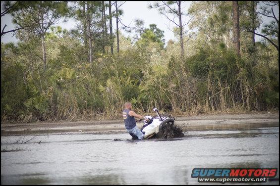 Man riding a white jet ski through a muddy puddle with trees in the background