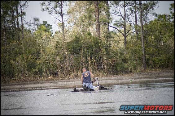 Man sitting on an ATV partially submerged in water with trees in the background