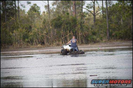 White ATV with a person riding it in muddy water, trees in background