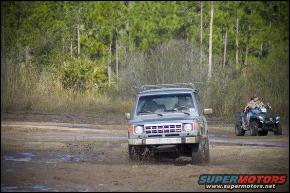 Dark green SUV with a roof rack driving through mud, front view. A person on an ATV is visible to the right
