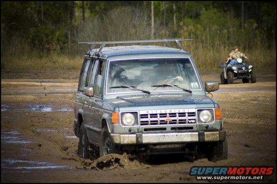 Blue Mitsubishi Pajero driving through mud, with an ATV in the background
