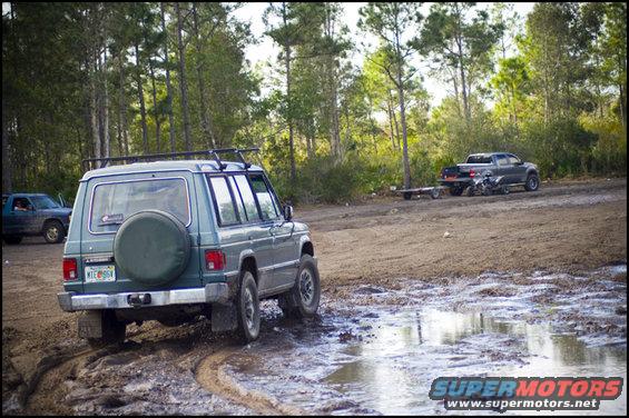 Green SUV with spare tire on back driving through mud, another truck with trailer in background