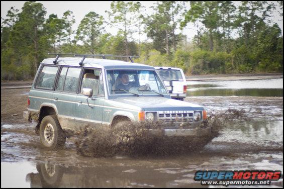 Green Mitsubishi Montero driving through mud, with a white van behind it