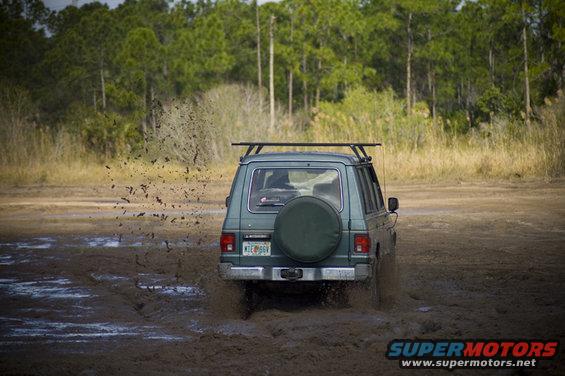 Green SUV with roof rack and spare tire on back driving through mud, kicking up spray