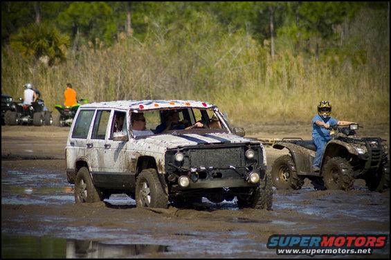 Mud-covered white Jeep Cherokee XJ with racing stripes on hood stuck in mud, another ATV nearby