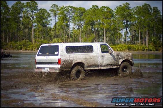 White Ford Bronco with camper shell driving through mud. Trees and water in background